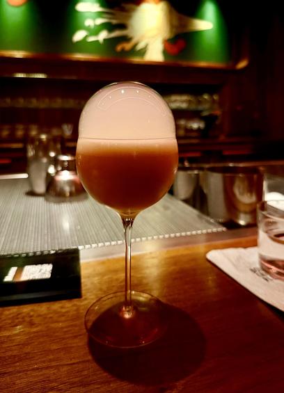 A color, medium-angle photograph in portrait orientation that captures a beautifully crafted cocktail known as the "Custard Bun" from Double Chicken Please cocktail bar on the lower east side of Manhattan. The drink is presented in an elegant, long-stemmed wine glass centered on a dark wooden bar top. The drink itself is an opaque, warm amber-tan color, topped with a prominent, translucent bubble dome that rests on the rim of the glass. The lighting is intimate and moody, typical of a high-end cocktail bar setting. The background is softly blurred, showing the warm, dimly lit interior of the bar. A dark green panel with faint, abstract decorative artwork is visible behind the bar, along with glimpses of stainless-steel bar equipment and glassware on a textured mat. To the right, a folded napkin and a small glass of water sit on the bar, while a menu rests to the left.
