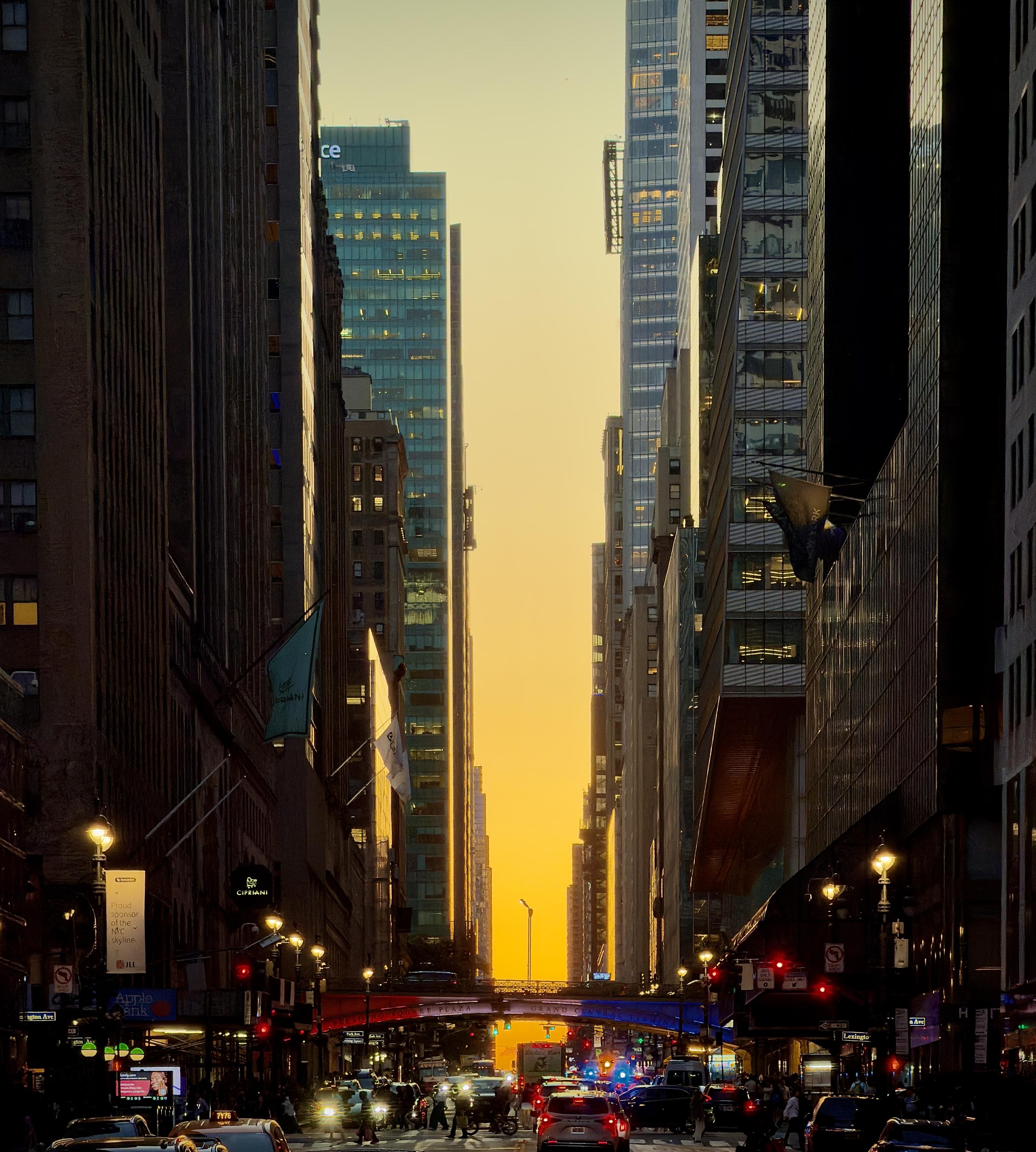 A color, wide-angle photograph in portrait orientation that captures a striking sunset illuminating 42nd Street, captured just east from the intersection with Lexington Avenue. The perspective looks down the urban canyon, where the brilliant, golden-orange sun is positioned perfectly at the end of the street, creating a vibrant, glowing pathway between the towering skyscrapers that flank both sides. The buildings, including notable structures near Grand Central Terminal, are cast in deep silhouette. The foreground is filled with the busy activity of a city street at dusk. Vehicles with their headlights and taillights glowing form a dense line of traffic, while pedestrians can be seen navigating the crosswalk. Above the street, decorative banners and signs are visible, and the iconic overpass near Grand Central is bathed in a red, white and blue light, contrasting with the warm, amber glow of the sunset.