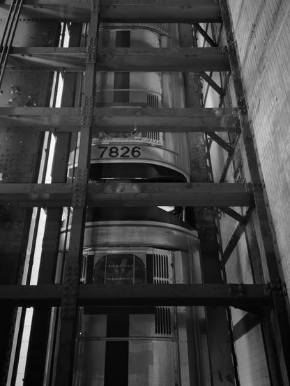 A noir, wide-angle photograph in portrait orientation taken inside of the Times Square–42nd Street subway station that captures a top-down perspective from an open, industrial stairwell looking directly onto a subway train passing through the station. The composition is heavily structured by the dark, rhythmic geometry of the iron staircase framing the scene, with thick steel beams and riveted supports creating a cage-like effect that looms over the tracks. The train car, marked with the bold number "7826" on its upper panel, fills the central vertical space. To the right, the wall of the station is lined with weathered, rectangular tiles that recede into the darkness.