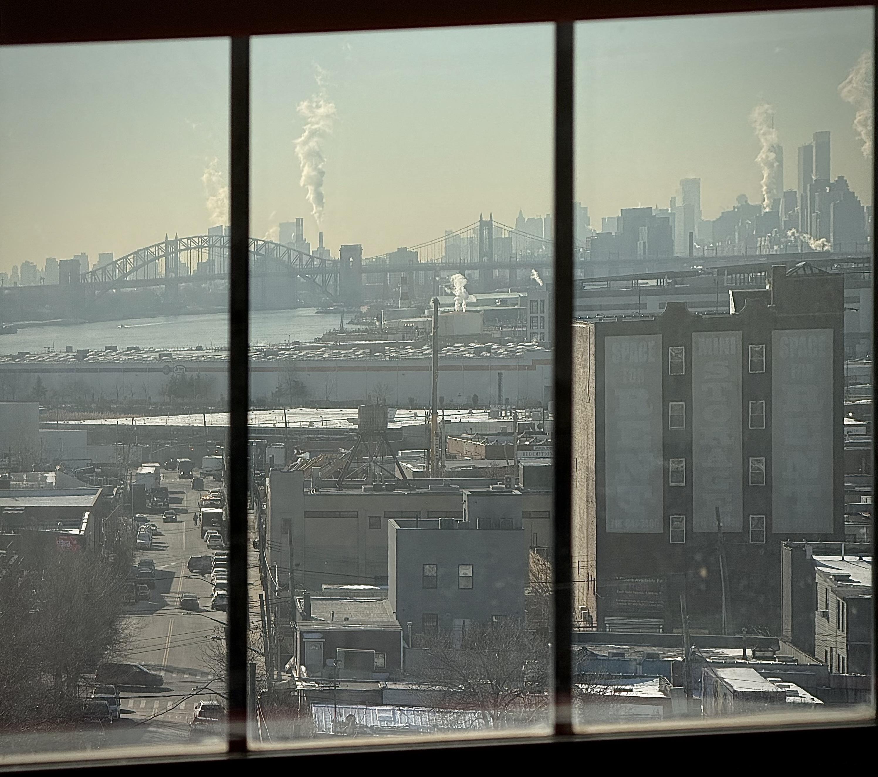 A color, wide-angle photograph in square aspect ratio that captures a view from an elevated vantage point, through a multi-paned window, looking out over an industrial area in the Hunts Point neighborhood of the Bronx, New York City. In the foreground, a multi-story, dark-colored building has large, weathered signage reading "SPACE FOR RENT" and "MINI STORAGE" on its side, overlooking a street where vehicles can be seen driving below. The immediate vicinity is filled with a dense mix of low-rise warehouses, industrial structures, and bare winter trees. In the distance is the East River, crossed by the arch of the Hell Gate Bridge. The skyline of Manhattan defines the horizon, visible through a soft, hazy atmosphere, punctuated by several plumes of white steam rising into the pale, bright sky