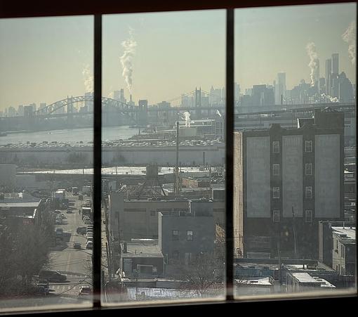 A color, wide-angle photograph in square aspect ratio that captures a view from an elevated vantage point, through a multi-paned window, looking out over an industrial area in the Hunts Point neighborhood of the Bronx, New York City. In the foreground, a multi-story, dark-colored building has large, weathered signage reading "SPACE FOR RENT" and "MINI STORAGE" on its side, overlooking a street where vehicles can be seen driving below. The immediate vicinity is filled with a dense mix of low-rise warehouses, industrial structures, and bare winter trees. In the distance is the East River, crossed by the arch of the Hell Gate Bridge. The skyline of Manhattan defines the horizon, visible through a soft, hazy atmosphere, punctuated by several plumes of white steam rising into the pale, bright sky