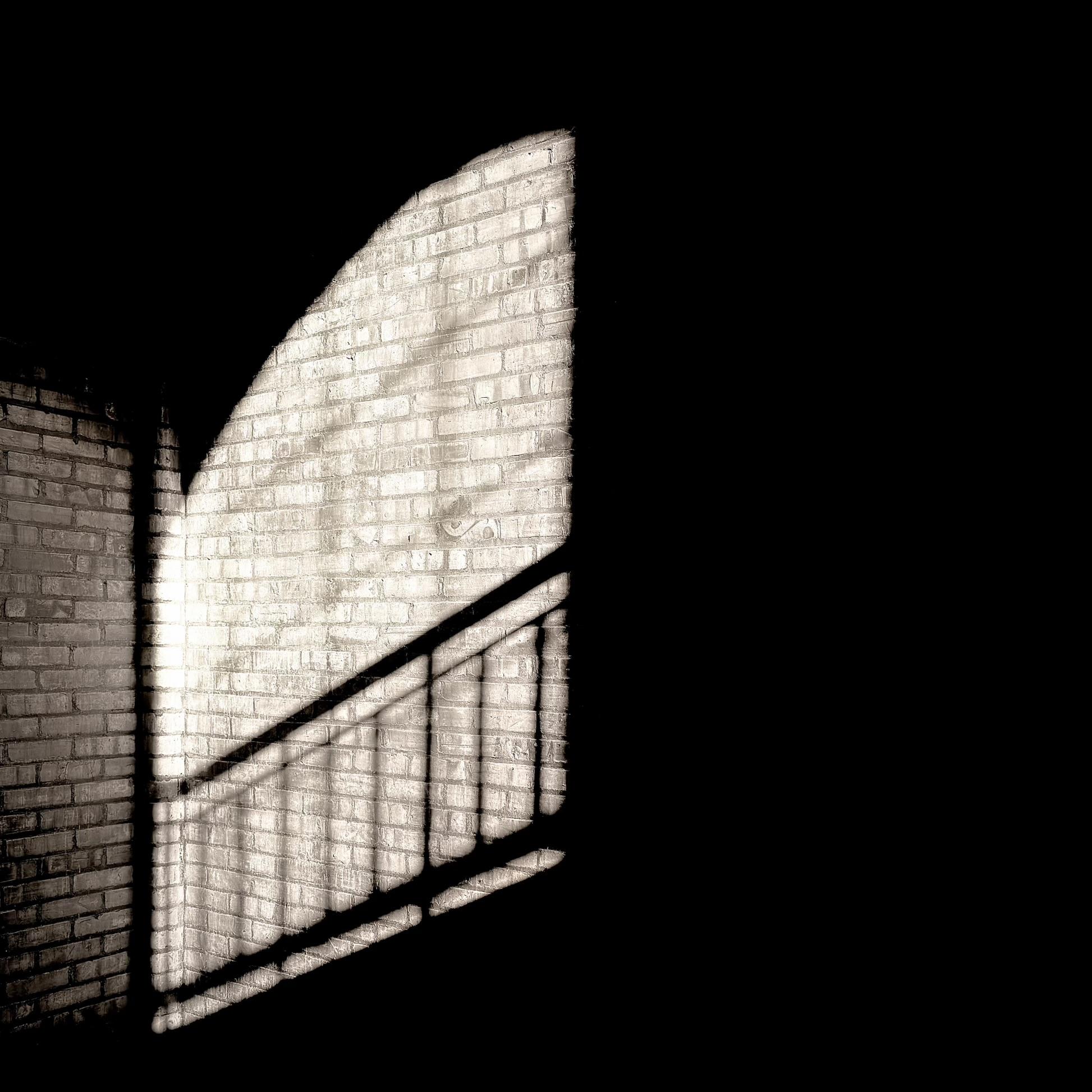 A desaturated color, wide-angle photograph in portrait orientation that captures a high-contrast scene in the stairwell of a warehouse in the Hunts Point neighborhood of the Bronx. It features a section of an interior brick wall illuminated by a sharp beam of sunlight cutting through darkness. The light reveals the textured, aged surface of the brickwork, which is oriented in a corner configuration where a side wall meets the main illuminated area. The sunlight casts a dramatic, elongated projection of a staircase railing onto the wall, with the dark shadows of the balusters creating a rhythmic, geometric pattern that ascends diagonally across the bricks. The window panes are also casting long shadows onto the wall.