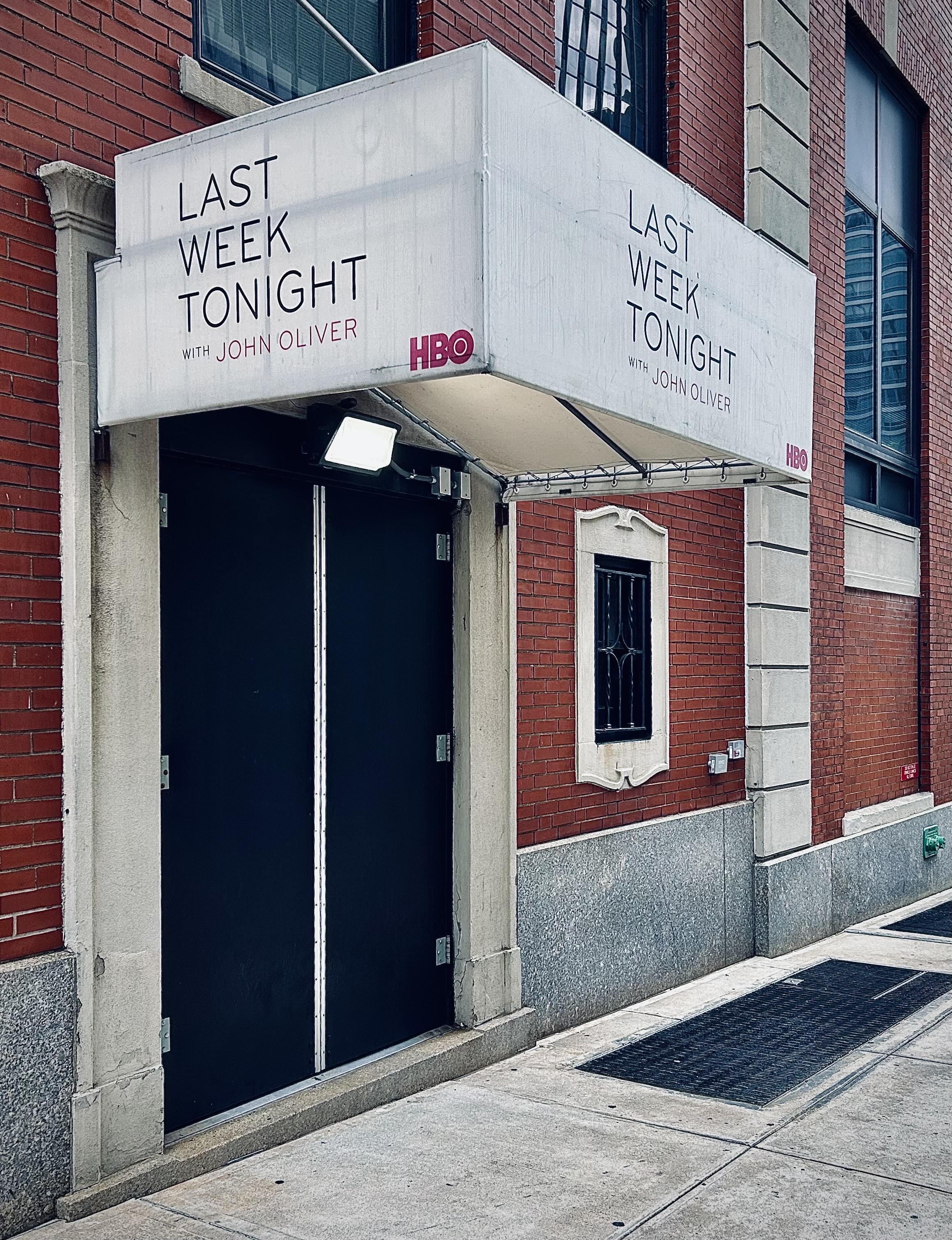 A color, wide-angle photograph in portrait orientation that captures a side entrance to the production site of "Last Week Tonight with John Oliver" in Manhattan. A prominent, white rectangular awning extends from a red brick wall, featuring the show's name printed in bold black letters with "WITH JOHN OLIVER" in a thinner, lighter font underneath. The HBO logo is displayed in the bottom right corner of the awning's front and side faces in a red color. Beneath the awning, a pair of tall, matte black double doors stands closed, framed by weathered, light-colored stone molding. To the right of the doors, a small, recessed window is visible, adorned with a decorative, ornate, and light-colored stone frame that echoes the design of the door frame. The exterior wall is constructed of classic red bricks, while a wide, light-colored stone base runs along the bottom of the building, separating it from the gray concrete sidewalk. A bright security light is mounted just under the edge of the awning, illuminating the upper portion of the doors. On the ground level, a large, rectangular metal grate is set into the pavement.