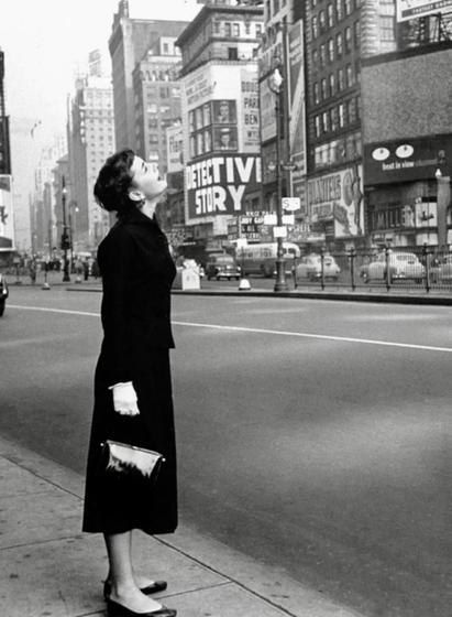 A noir, wide-angle photograph in portrait orientation that features a young Audrey Hepburn standing on a sidewalk in Times Square, Manhattan, in October of 1951. Captured in profile, she is dressed in a dark, tailored coat and skirt, wearing light-colored gloves and holding a small, dark clutch bag. Hepburn is positioned on the left side of the frame, gazing upward with an expression of wonder or contemplation, her slender silhouette starkly contrasted against the vast, empty street stretching out behind her. The background captures the dense, urban landscape of mid-century New York City, characterized by towering buildings covered in large-scale advertisements and signage, including a prominent display for the film Detective Story. The street itself is relatively quiet, with a few vintage automobiles visible in the distance, emphasizing the isolation of the subject.