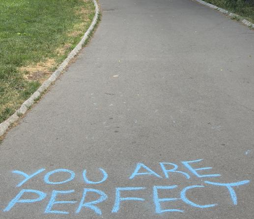 A color, wide-angle photograph in square aspect ratio taken in Central Park, New York City, that captures an asphalt walkway curving slightly toward the upper portion of the frame. Written directly onto the foreground of the pavement in bright, light blue chalk is the encouraging message, "YOU ARE PERFECT". The block lettering is capitalized, large, and hand-drawn, spanning almost the entire width of the path. The asphalt itself is textured and gray, showing slight wear with a few faint, scattered chalk marks, a stray twig, and a small leaf further up the path. To the left of the asphalt path, a natural stone curb separates the pavement from a lush green lawn, which is dotted with tiny white clover flowers. The path curves away from the viewer, guiding the eye from the vibrant blue text in the foreground toward the walkway ahead. The lighting from the overcast afternoon sky is soft and even.