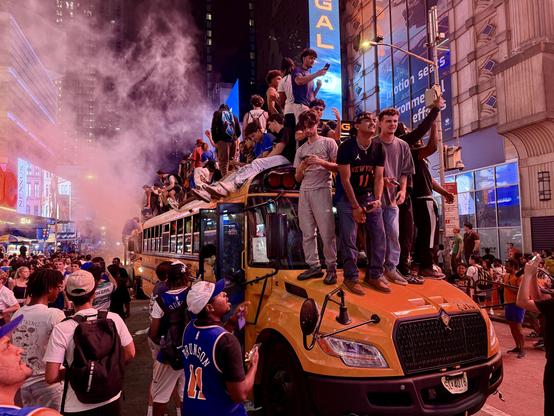 A color, wide-angle photograph taken at night in the Times Square neighborhood of Manhattan after the Knicks won the NBA finals. A chaotic and ecstatic crowd celebrates in the packed street. The central focus is a yellow school bus completely covered with young men who have climbed onto its hood, front bumper, and entire roof to celebrate. A thick cloud of pinkish-red smoke billsows from behind the bus, lit up by the bright billboards. On the hood of the bus, three young men stand triumphantly; the one in the middle wears a New York Knicks jersey with the number 11. Behind them, dozens of other fans are crammed onto the roof of the bus, some holding up their phones to record the moment, while others sit along the edge or stand precariously above the crowd. On the street level surrounding the bus, a dense sea of people fills the frame, creating an intense, high-energy block party atmosphere. In the foreground, spectators look up at the bus, including a man facing away from the camera wearing a blue Knicks jersey with "BRUNSON 11" printed on the back. The glowing blue and red lights of Times Square skyscrapers tower in the background, adding a cinematic glow to the scene.