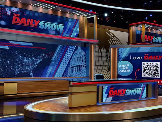 A color, wide-angle photograph that captures the brightly lit, modern television studio set of The Daily Show on Comedy Central, photographed from at low angle from in front of the audience. The focal point of the set is the main anchor desk, which rests on an elevated, oval-shaped wooden platform trimmed with a glowing white LED light strip. The desk itself is crafted from polished wood panels with tan accents, a reflective dark glass top, and a built-in rectangular screen on its front displaying the show's red, white, and blue logo. Behind the empty black office chair sits an expansive, multi-layered backdrop consisting of wood framing and large digital screens. The primary screen on the left displays a stylized, dark blue map of North America alongside a graphic of the United States Capitol dome. To the right side of the frame, the set incorporates a distinct promotional screen that reads "Love DAILY" and features a prominent QR code next to a teal circle with the text "GRAB THE GEAR!". A red arrow points directly to the QR code, encouraging audience interaction. In the background between the screen structures, a large graphic of a world globe outline is visible on a neutral-toned wall. The overhead studio environment is visible at the very top, showcasing professional grid lighting, rigs, and small red emergency exit signs, all casting a polished, high-production glow over the glossy dark floor of the studio.