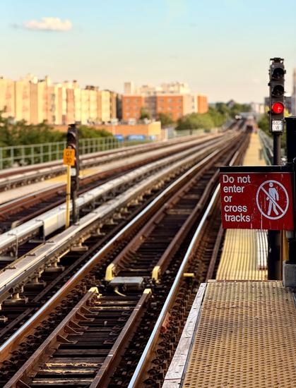 A color, wide-angle photograph in portrait orientation that captures a ground-level perspective from the edge of the outdoor elevated subway platform at Pelham Parkway station, looking down a long, straight stretch of parallel train tracks. The foreground prominently features a yellow, textured tactile platform edge on the right, above which a weathered red warning sign reads, "Do not enter or cross tracks," accompanied by a white graphic of a person with a slash through it. Just behind the sign, a tall, black metal signal post stands with a red light illuminated. The track system stretches forward into a deep linear perspective, with the metal rails, wooden ties, and gravel beds creating a strong sense of depth as they converge toward the distant background. On the left side of the tracks, a smaller, yellow rectangular sign labeled "GT 30" is mounted on a short post next to a secondary track signal. The middle and background are softly blurred due to a shallow depth of field, highlighting the cityscape under a clear blue sky with a few light clouds. Low-rise residential buildings in shades of tan and brick-red line the left side of the tracks, partially obscured by green treetops, while a faint silhouette of a train can be seen far off in the distance where the tracks begin to curve. The scene is bathed in warm, natural sunlight, casting soft shadows.