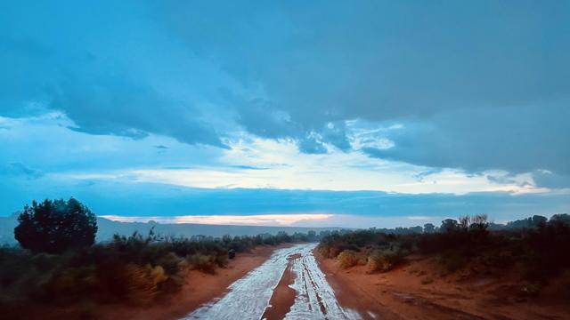 A color, wide-angle photograph in landscape orientation that captures a desert scene in Coconino County, Arizona. A wet dirt road cuts through a vast, rural desert landscape immediately following a heavy downpour. The unpaved road, rich with red-orange mud, stretches from the foreground toward a distant, flat horizon line. Its surface is deeply saturated, covered in wet, reflective tire tracks and shallow pools of standing water that mirror the bright sky above, giving the track a gleaming, silver-white appearance down its center. Low-lying, dark green and brownish desert shrubs and scattered vegetation line both sides of the road, rooted in the damp, red earth, with a single larger, dark green tree standing out on the far-left side of the frame. Dominating the upper two-thirds of the image is an expansive, dramatic sky filled with thick, low-hanging storm clouds. The sky is rendered in a deep, moody palette of blues, teals, and dark grays, conveying a cool, atmospheric quality. A sliver of clear sky breaks along the distant horizon, casting a soft, pale white and subtle yellowish-pink glow that hints at sunset, contrasting sharply with the heavy, dark cloud cover above.