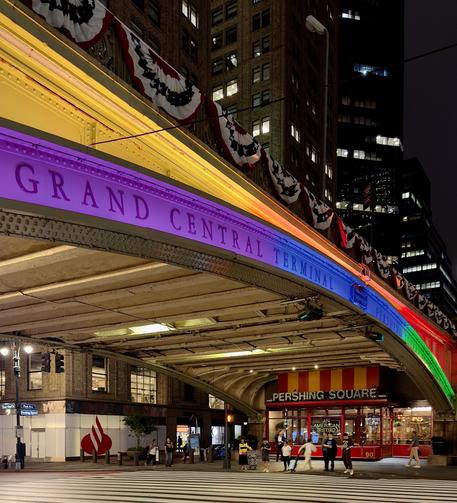 A color, wide-angle photograph in square aspect ratio that captures the nighttime scene outside of Grand Central Terminal facing the Pershing Square Viaduct bridging across 42nd Street. The prominent steel overpass curves from the upper left toward the middle right of the frame, emblazoned with the words "GRAND CENTRAL TERMINAL" in all-caps lettering. For Pride month, the bridge is vividly illuminated by a spectrum of neon rainbow lighting that shifts from purple and violet on the left, to blue, green, and a bright red-orange gradient toward the right. Red, white, and blue patriotic bunting hangs in decorative swags along the upper railing of the viaduct, contrasting with the colorful lighting underneath. Below the illuminated bridge, a crosswalk with bold white stripes stretches across the foreground. On the far side of the street, tucked directly beneath the overpass, is the brightly lit exterior of the "Pershing Square" restaurant, featuring a red and yellow striped awning and a vibrant red facade with large glass windows. Several pedestrians are scattered along the sidewalk and near the restaurant entrance, some walking or standing in small groups. To the left, a street corner features a storefront with a large red Santander bank logo, while towering, dark skyscrapers with glowing office windows rise into the night sky in the background, framing the scene.