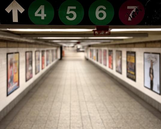 A color, wide-angle photograph in square aspect ratio that captures an eye-level perspective looking down a long, empty subway corridor at Grand Central Terminal in New York City. In the immediate foreground, a black overhead directional sign stretches horizontally across the top of the frame. The sign features a white arrow pointing straight ahead, followed by subway line emblems: three green circles containing the numbers "4", "5", and "6" respectively, and a purple circle containing the number "7" with a slightly weathered, scratched surface. Just below this sign, the corridor extends deep into the background, narrowing toward a central vanishing point. The focus of the camera is sharply fixed on the overhead signage, while the rest of the hallway softly blurs as it recedes into the distance, emphasizing a sense of depth and quiet emptiness. The walls of the corridor are lined with rectangular white tiles, bordered along the bottom by a dark grey base. Spaced evenly along both the left and right walls are framed, colorful advertisements that add pops of color to the otherwise muted beige, white, and grey palette of the walkway. The floor is composed of light-colored, gridded tiles that reflect the soft, overhead fluorescent lighting strips running down the length of the ceiling. The tunnel is completely devoid of people, evoking a serene and unusually still atmosphere within a typically bustling transit hub.
