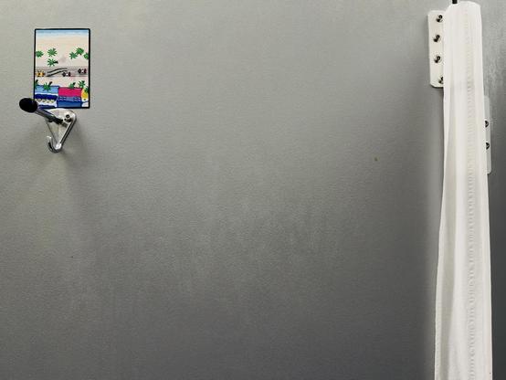 A color, medium-angle photograph in landscape orientation that captures a close-up view inside a public restroom stall at a Target retail store, focusing on the gray partition door. On the upper-left side of the wall, a shiny chrome coat hook with a black rubber tip is securely mounted with screws. Placed directly above this hook is a small, vertically oriented rectangular sticker featuring pixelated, retro-style artwork. The sticker depicts a l coastal scene with a yellow sports car driving along a grey road, leaving tire tracks in its path as it is being chased by two police cars. The background of the artwork includes sand, green palm trees, a bright blue ocean, and colorful buildings, contrasting sharply with the plain, neutral tones of the partition. The rest of the composition emphasizes the stark, utilitarian environment of the restroom stall. The middle and lower sections of the image are dominated by the smooth, matte gray surface of the stall divider, which shows subtle variations in light and texture. On the far right edge, a white vertical strip of toilet paper is visible, attached to the gap in the door above a metal bracket. The lighting is even and bright, casting minimal shadows.