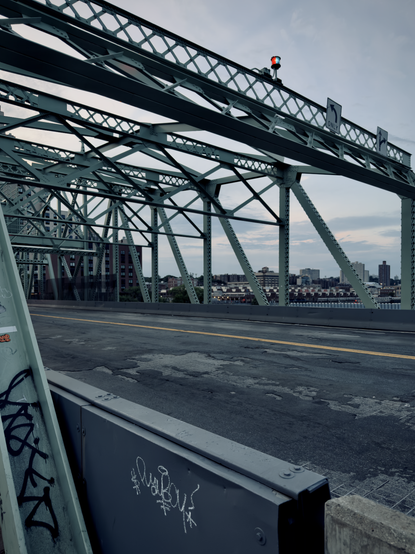 A color, wide-angle photograph in portrait orientation that captures a low-angle view looking down an empty, weathered roadway on the University Heights Bridge, facing toward the Inwood neighborhood of Manhattan near sunset. The foreground is dominated by the intricate, pale green painted steel truss framework of the bridge, which arches over the asphalt road. The road surface shows significant wear and patching, marked with a double yellow line running down the center. In the immediate foreground, a grey metal barrier and the green steel support beam are tagged with black and white graffiti. Atop the overhead steel crossbeam, traffic signs indicating turn lanes are visible alongside a small red and blue signal light. Through the gaps in the bridge's massive geometric steel lattice, the urban landscape of Inwood is visible in the midground across the water. A mix of multi-story residential and commercial buildings, including a prominent brick building on the left, lines the distant shore under a pale, partly cloudy sky. The lighting late evening, with soft, muted tones of blue, grey, and hints of a faint warm glow on the horizon from the setting sun.