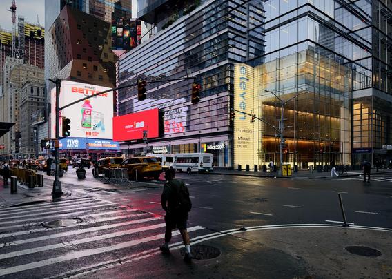 A color, wide-angle photograph in landscape orientation that captures a street-level view of Eighth Avenue near Times Square, Manhattan on a damp, overcast morning, showcasing the wet asphalt reflecting vibrant city lights. In the foreground, a pedestrian wearing shorts, a dark jacket, and a backpack crosses a wet, striped crosswalk, moving away from the viewer. The slick asphalt street catches the red and white glow from the massive, bright electronic billboards lining the buildings in the background. Yellow cabs, a white commuter shuttle, and other vehicles are stopped or moving slowly through the intersection under traffic lights glowing red. Dominating the right side of the scene is a modern high-rise skyscraper with a glass-facade lobby. A vertical glowing yellow sign on the building reads "eleven," with tenant logos for companies like Microsoft and Proskauer displayed below it. Further down the street on the left, massive digital billboards advertise brands like Gatorade and Verizon, casting a powerful pink and white light over the scene. In the far background, additional skyscrapers rise into the hazy sky, including one under construction with a crane, emphasizing the dense, vertical architecture of the New York City landscape.