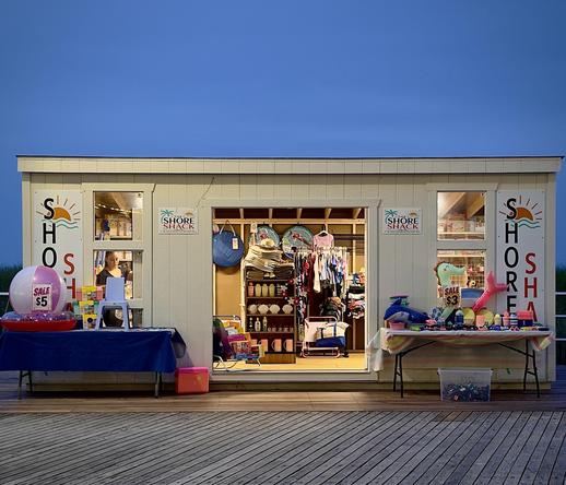 A color, wide-angle photograph in square aspect ratio that captures the scene along the boardwalk in Atlantic City. The focal point of the scene is an eye-level view of the "Shore Shack," a rectangular, cream-colored wooden kiosk illuminated from within against a deep, solid blue twilight sky. The shop stands on a wooden boardwalk made of diagonal planks that fill the lower third of the frame. The front of the kiosk features a large central open doorway revealing an organized interior packed with summer merchandise.. Flanking the entrance are two vertical white banners featuring the store's name, "SHORE SHACK," written stacked vertically alongside a logo of a stylized sun setting over waves and a palm tree. To the left, a person is partially visible inside a window booth, while a blue-cloth-covered folding table out front displays a large pink and white inflatable beach ball with a "SALE $5" sign and stacked colorful items. To the right, a second folding table covered in a striped pastel cloth displays an assortment of pool and beach toys. The warm, bright indoor lighting spills out onto the immediate boardwalk area, casting a soft glow that contrasts sharply with the smooth, unclouded gradient of the evening sky.