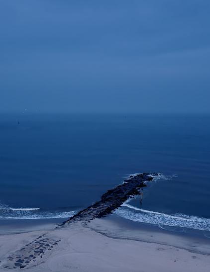 A color, wide-angle photograph that captures an elevated, high-angle view of a beach and the ocean in Atlantic City, New Jersey during twilight, dominated by a deep, monochromatic blue color palette. In the foreground, a sandy shore stretches across the bottom of the frame, with a line of dark paving stones visible on the left side of the beach. A long, narrow rock jetty constructed from dark boulders juts straight out from the shoreline into the sea. Small, white-foamed waves lap gently against the sides of the jetty and along the sandy shoreline. The vast ocean occupies the middle section of the image, transitioning seamlessly into a heavy, overcast sky at a faint, hazy horizon line. The entire scene is enveloped in the cool, blue-gray light of dusk. Far out on the left side of the horizon, a couple of tiny, faint green lights are visible, indicating distant ships.