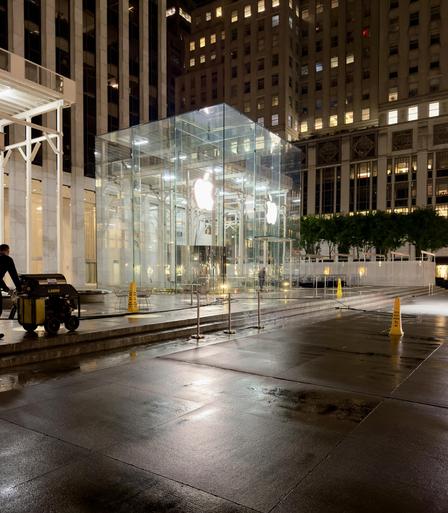 A color, wide-angle photograph in portrait orientation that captures the glass cube of the Fifth Avenue Apple Store standing brightly illuminated against the night sky, now fully uncovered. The glowing white Apple logo is clearly visible through the transparent glass panels, which reflect both the interior fixtures and the surrounding city lights. The foreground features a wide, expansive plaza with wet, dark pavement that glistens from recent cleaning. On the far left, a worker is captured from behind, operating a black and yellow mobile pressure washer. Multiple bright yellow caution cones are placed around the damp plaza, positioned near metal stanchions, marking off the area during the early morning cleanup process. The background is dominated by towering, classic skyscrapers with grid-like patterns of lit windows, framing the plaza in a dense urban setting. To the left of the glass cube, a few remaining scaffolding columns stand near the building's base. On the right side, a row of manicured green trees is visible behind a long, white temporary barrier.