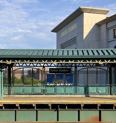 A color, wide-angle photograph in square aspect ratio that captures a bright, clear morning light illuminating the 161st Street–Yankee Stadium subway station in the Bronx. In the foreground, a prominent elevated subway platform structure features a green, corrugated metal roof supported by matching green metal pillars and intricate, decorative ironwork railings. Centered on the platform, a dark sign with white lettering clearly reads "Yankee Stadium," situated above blue advertisements for Spectrum Television mounted between the metal railings. The platform floor is concrete with a yellow tactile warning strip running along the edge, while the support structure below the tracks is also painted in a weathered green finish. Behind the station platform, the massive, light-colored stone facade of Yankee Stadium rises against a vibrant blue sky filled with wispy, scattered clouds. The stadium name is etched in gold lettering near the top of the building's exterior, which is bathed in warm, directional morning sunlight. In the distance, visible through the station's iron lattice, a cluster of tall, brown apartment buildings stands above a patch of green trees, providing a sense of scale and context to the scene.