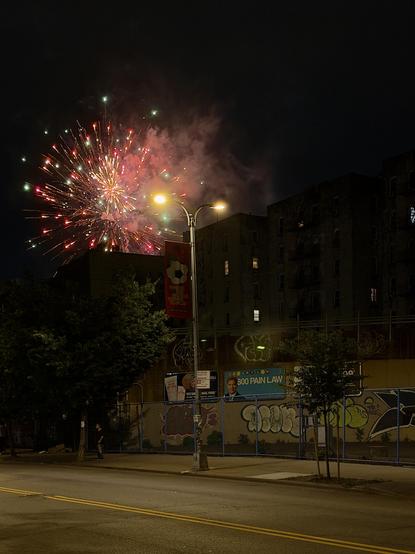 A color, wide-angle photograph in portrait orientation that captures a vibrant aerial firework display illuminating the night sky behind a row of multi-story apartment buildings in New York City. The burst of light is predominantly composed of red, gold, and green sparks, radiating outward against the dark backdrop with a lingering haze of smoke trailing from the explosion. The urban setting is defined by several dark, brick residential buildings that frame the firework, with lit windows that contrast with the deep shadows of the evening. In the foreground, an empty city street is lined with a sidewalk, a chain-link fence, and a partitioning wall covered in extensive graffiti. A tall, double-headed lamppost stands prominently in the center, featuring a red banner hanging from it that displays a soccer ball graphic and text. To the left and right, sparse trees stand along the curb, and billboard advertisements for a law firm are visible along the lower wall near the sidewalk. A person stands in dark clothing at the base of one of the trees directly beneath the explosion in the sky.