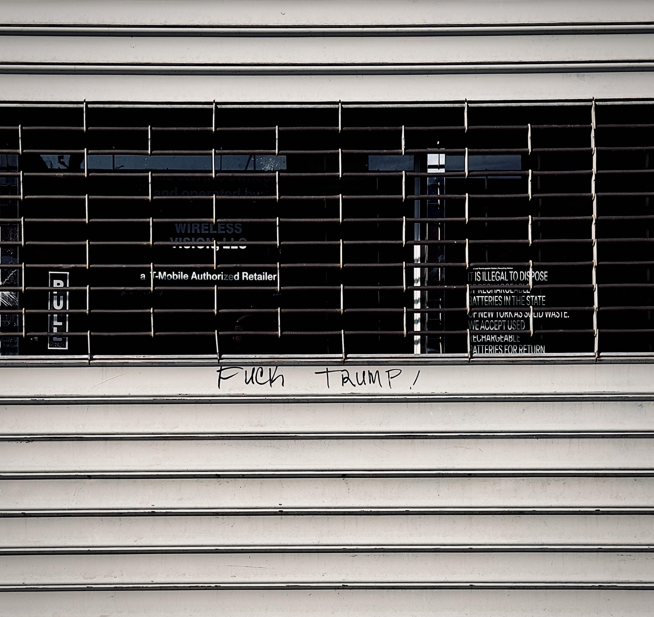 A color, wide-angle photograph in square aspect ratio that captures a closed T-Mobile storefront, shielded by a metal rolling security gate that is pulled down. The gate features a horizontal barred window section at eye level, revealing the dark, obscured interior of the retail store behind the glass. Through the bars, signage for "WIRELESS VISION, LLC" and "a T-Mobile Authorized Retailer" is visible on the interior glass, along with a notice stating that it is "illegal to dispose" of batteries in the state. On the flat surface of the gate, directly below the barred section, the phrase "FUCK TRUMP!" is written in black marker. The lettering is simple and slightly uneven, appearing as a piece of hand-written graffiti centered on the lower portion of the gate. The surrounding metal shutter is horizontally ribbed, adding texture to the otherwise plain, light-colored surface. Morning sunlight is evenly illuminating the scene.
