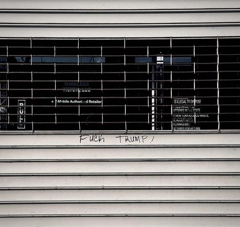 A color, wide-angle photograph in square aspect ratio that captures a closed T-Mobile storefront, shielded by a metal rolling security gate that is pulled down. The gate features a horizontal barred window section at eye level, revealing the dark, obscured interior of the retail store behind the glass. Through the bars, signage for "WIRELESS VISION, LLC" and "a T-Mobile Authorized Retailer" is visible on the interior glass, along with a notice stating that it is "illegal to dispose" of batteries in the state. On the flat surface of the gate, directly below the barred section, the phrase "FUCK TRUMP!" is written in black marker. The lettering is simple and slightly uneven, appearing as a piece of hand-written graffiti centered on the lower portion of the gate. The surrounding metal shutter is horizontally ribbed, adding texture to the otherwise plain, light-colored surface. Morning sunlight is evenly illuminating the scene.