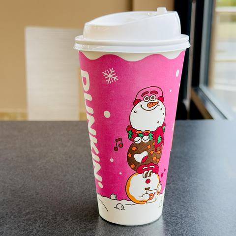 A color, medium-angle photograph in square aspect ratio that captures a large-sized paper Dunkin’ Donuts coffee cup sitting on a dark grey table inside of the coffee shop. The cup is unseasonably adorned with a bright, cheerful magenta-pink background featuring a playful, winter-themed illustration. The design prominently features a vertical stack of three cute, cartoon-like characters: a white snowman wearing a pink hat at the top, a brown donut-shaped character in the middle, and a white, round character wearing a red hat at the bottom holding a small cup. A red scarf decorated with small green pine trees wraps around the snowman and the donut, while musical notes float nearby. The top of the cup is fitted with a clean, white plastic lid with a sip opening. Running vertically down the left side of the cup, the word "DUNKIN" is printed in large, bold, white, capital letters. The background of the cup is lightly decorated with white snowflakes and small circular dots, mimicking falling snow, while the bottom of the cup features a white, wavy line representing snow on the ground. The morning lighting is bright and even.