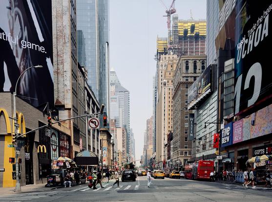 A color, wide-angle photograph in landscape orientation that captures a street-level view of Eighth Avenue near Times Square in New York City, dominated by towering glass skyscrapers and prominent digital billboards. On the left, a large McDonald’s sign stands out, while various advertisements, including a "Kalshi" promotion featuring the name "OCHOA," appear on large screens and building facades. The street is populated by a few pedestrians crossing the road, a parked black car, and a bright red double-decker "TOPVIEW" tour bus. A construction crane is visible in the background, hovering over another building. The atmosphere in the image is noticeably hazy and muted. The smoke from wildfires has created an unhealthy environment, obscuring the clarity of the cityscape and casting a dull, washed-out quality over the buildings and sky.