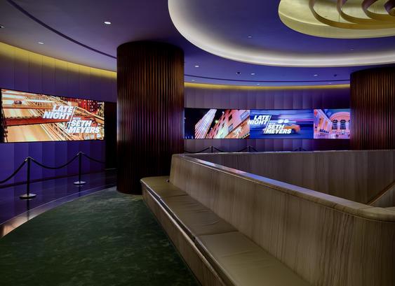 A color, wide-angle photograph in landscape orientation that captures the modern, curved interior hallway outside the Late Night with Seth Meyers studio at NBC Studios in Rockefeller Center. A long, cushioned bench with a light-wood exterior follows the gentle curve of the hallway, sitting atop a deep green carpet. The walls are panelled and feature large, rectangular digital displays showing the show’s title card, Late Night with Seth Meyers, superimposed over vibrant, blurred images of New York City landmarks like the Brooklyn Bridge and Grand Central Terminal. The area is defined by dark, ribbed cylindrical pillars that provide a strong vertical contrast to the sweeping horizontal lines of the architecture. The lighting is soft and atmospheric, with recessed ceiling lights casting a warm, ambient glow that highlights the polished textures of the wood and the clean, contemporary design of the space. Black stanchions with connecting velvet ropes stand before the screens.