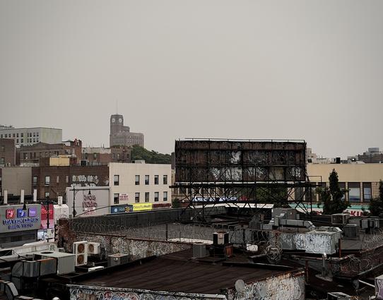 A color, wide-angle photograph in landscape orientation taken from an elevated transit platform that captures the urban expanse of the Fordham neighborhood in the Bronx under a thick, monochromatic haze caused by wildfire smoke. The sky is an oppressive, featureless flat gray, draining the scene of color and softening the details of the distant skyline. Dominating the middle-ground skyline is the prominent, historic Bronx Borough Courthouse, its iconic clock tower rising above the mid-rise brick residential and commercial buildings that stretch across the horizon. In the foreground, the rooftops of the local commercial strip are cluttered with industrial details, including weathered HVAC units, sprawling vent ductwork, and layers of graffiti that adorn both the low brick walls and the roof structures. A large, dark, skeletal metal billboard frame stands starkly against the hazy background, its structure partially obscured by tangled graffiti. Coils of razor wire run along the edges of the roof parapets. 