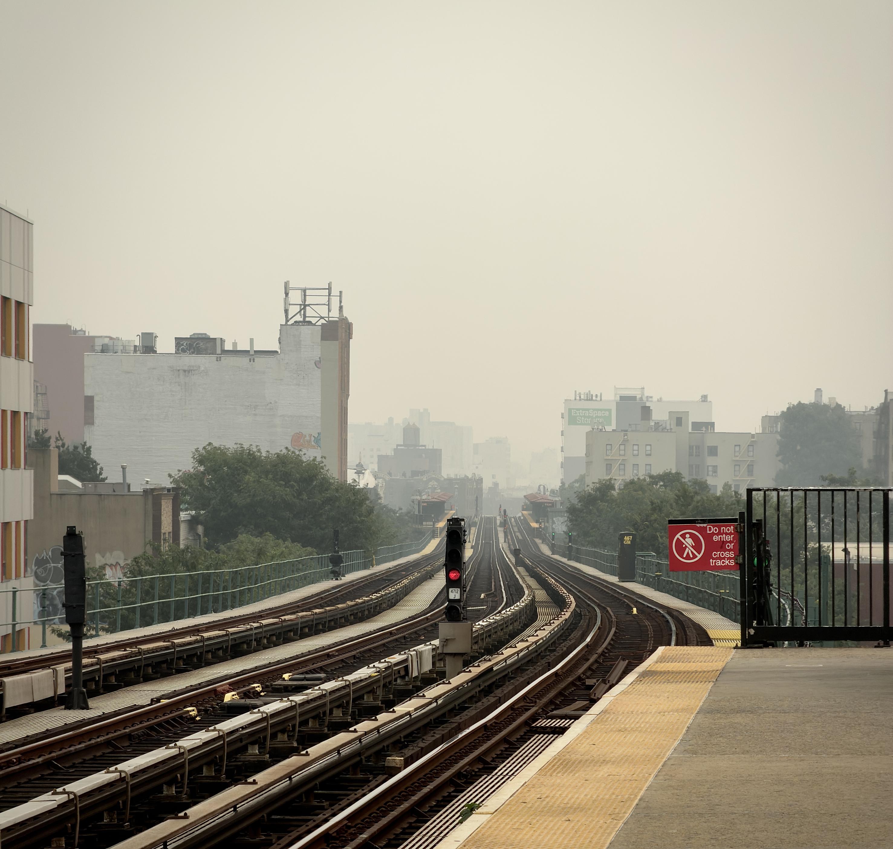 A color, wide-angle photograph in square aspect ratio captured from the elevated subway platform at the Fordham Road Station in the Bronx, looking south towards Manhattan under a thick, monochromatic haze caused by wildfires. In the foreground, the concrete subway platform edge features a yellow tactile warning strip, and a prominent red sign on a black metal fence warns, "Do not enter or cross tracks." The train tracks stretch into the distance, flanked by a signal pole with a glowing red light situated between the tracks. The sky is an oppressive, hazy gray, completely obscuring any distant landmarks like the Empire State Building that would typically be visible in this sightline. The surrounding urban landscape is muted by the thick atmosphere, creating a deep sense of depth as buildings fade into the dense, foggy horizon. To the left, a tall, nondescript building with some graffiti at its base borders the tracks, while various mid-rise residential and commercial structures are visible in the background, their details softened by the heavy air. Patches of green trees peek through between the buildings, and additional signal lights and infrastructure markers line the tracks, leading the eye toward the center of the composition where the tracks converge into the hazy, indistinct distance.