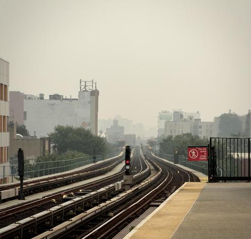A color, wide-angle photograph in square aspect ratio captured from the elevated subway platform at the Fordham Road Station in the Bronx, looking south towards Manhattan under a thick, monochromatic haze caused by wildfires. In the foreground, the concrete subway platform edge features a yellow tactile warning strip, and a prominent red sign on a black metal fence warns, "Do not enter or cross tracks." The train tracks stretch into the distance, flanked by a signal pole with a glowing red light situated between the tracks. The sky is an oppressive, hazy gray, completely obscuring any distant landmarks like the Empire State Building that would typically be visible in this sightline. The surrounding urban landscape is muted by the thick atmosphere, creating a deep sense of depth as buildings fade into the dense, foggy horizon. To the left, a tall, nondescript building with some graffiti at its base borders the tracks, while various mid-rise residential and commercial structures are visible in the background, their details softened by the heavy air. Patches of green trees peek through between the buildings, and additional signal lights and infrastructure markers line the tracks, leading the eye toward the center of the composition where the tracks converge into the hazy, indistinct distance.