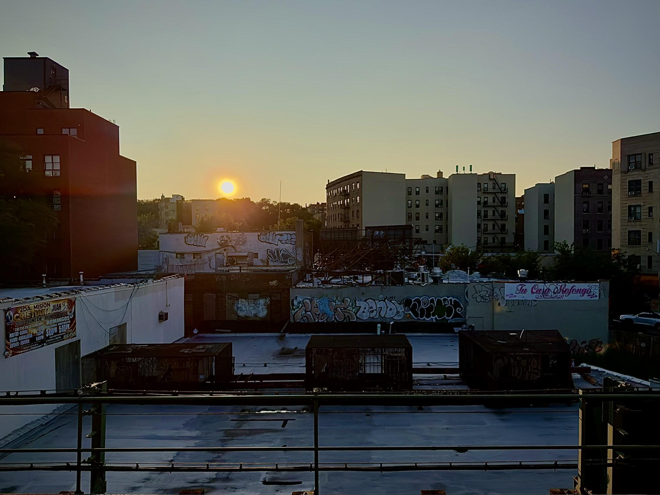 A color, wide-angle photograph in landscape orientation that captures an eye-level shot from an elevated subway platform captures a cityscape view during a bright sunset, framed by a dark metal railing at the bottom edge. In the foreground, a large, flat rooftop covered in pale material features several boxy, dark structures and is bordered by white and brick-faced walls covered in extensive graffiti. The sun hangs low in the center-left horizon, glowing as a small, intense orb against a pale, clear evening sky. Mid-rise apartment buildings and commercial structures stretch across the background, showing a mix of architectural styles, including a tall red-brick building on the far left and beige apartment blocks with external fire escapes on the right. The lighting casts a soft, warm glow over the urban scene, highlighting the varied textures of the rooftops, graffiti tags, and windows of the distant buildings.