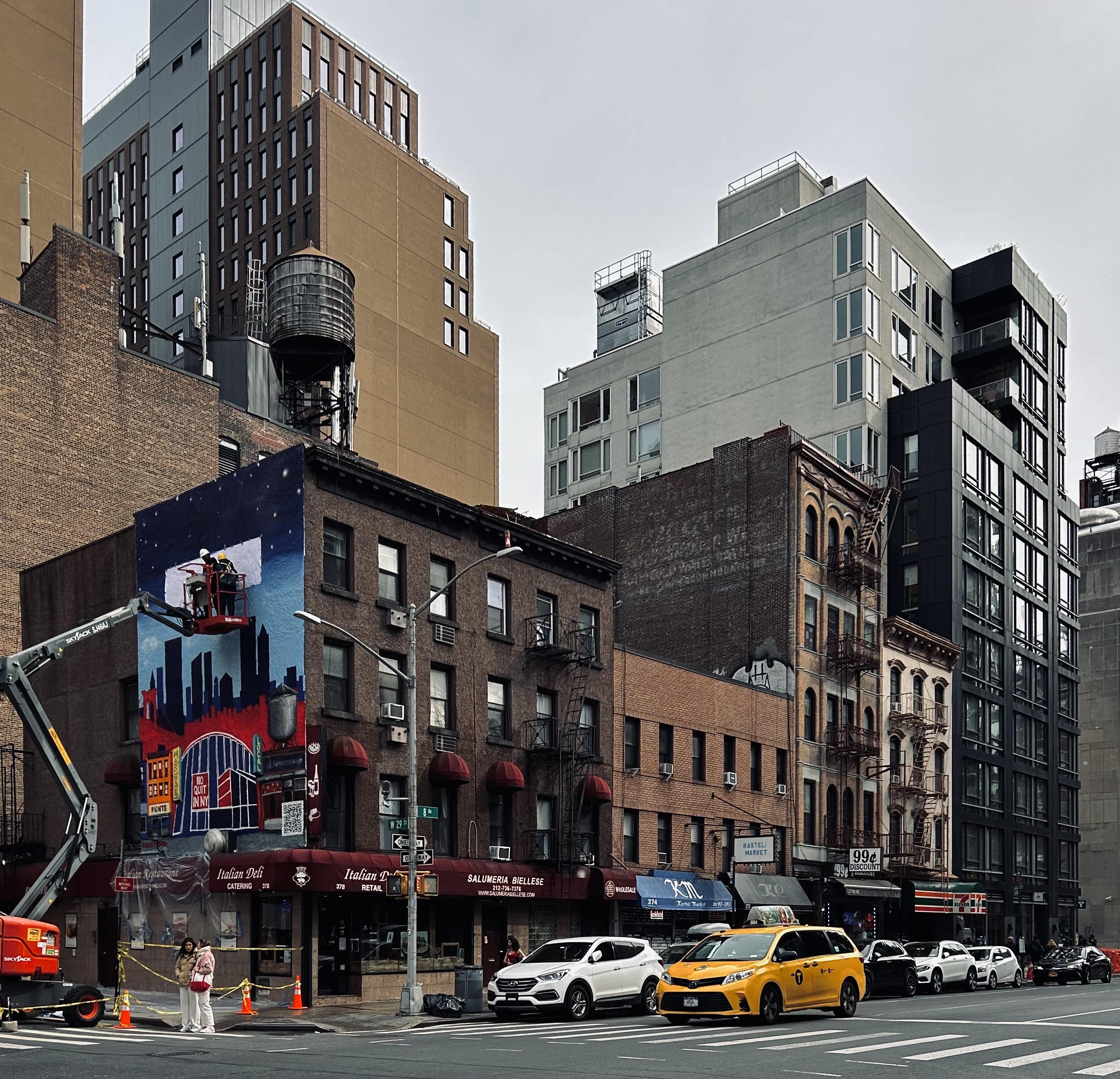A color, wide-angle photograph in landscape orientation that captures a bustling corner in the Chelsea neighborhood of Manhattan under an overcast sky. In the foreground, a vibrant, multi-story mural is being painted on the side of a classic brick building by two workers in a cherry picker. The mural features a stylized skyline with deep blues, reds, and blacks. At the base of this building sits "Salumeria Biellese," an Italian deli with a prominent red awning. A yellow New York City taxi and several white cars are positioned in the street, while two pedestrians stand near yellow caution tape and road cones on the sidewalk. Modern high-rise apartments and older industrial-style buildings with fire escapes and rooftop water towers create a layered architectural backdrop. The composition emphasizes the intersection of old and new New York. The central brick building, with its traditional red awnings and gridded windows, is flanked by a sleek, contemporary grey and black glass residential tower to the right and a tall, tan building behind it. To the right of the deli, a row of smaller storefronts includes a "99¢ Discount" shop and a 7-Eleven. Street infrastructure, such as silver lamp posts, traffic signals, and a crosswalk, anchors the scene in an active urban environment. The lighting is soft and even, highlighting the textures of the brickwork, the metallic sheen of the construction equipment, and the vivid colors of the developing mural.