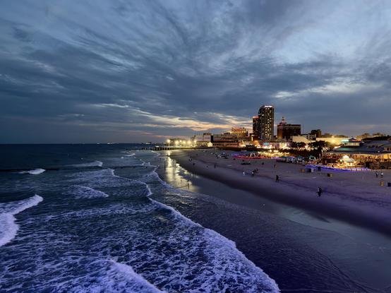 A color, wide-angle photograph in landscape orientation that captures an elevated view of the Atlantic City coastline at dusk under a dramatic, swirling sky filled with moody blue and purple clouds. Gentle waves with white foam roll onto a wide, sloping sandy beach on the right, while the dark, calm ocean stretches out to the horizon on the left. The fading light of sunset creates a faint, warm glow on the distant horizon line just above the water. Along the beachfront, the city skyline lights up with glowing high-rise hotels, casinos, and string-lit establishments. Small figures of people can be seen walking along the damp sand near the shoreline and gathered around illuminated beach bars and structures. The warm artificial lights contrast sharply with the cool, twilight tones cast across the water and the expansive evening sky.