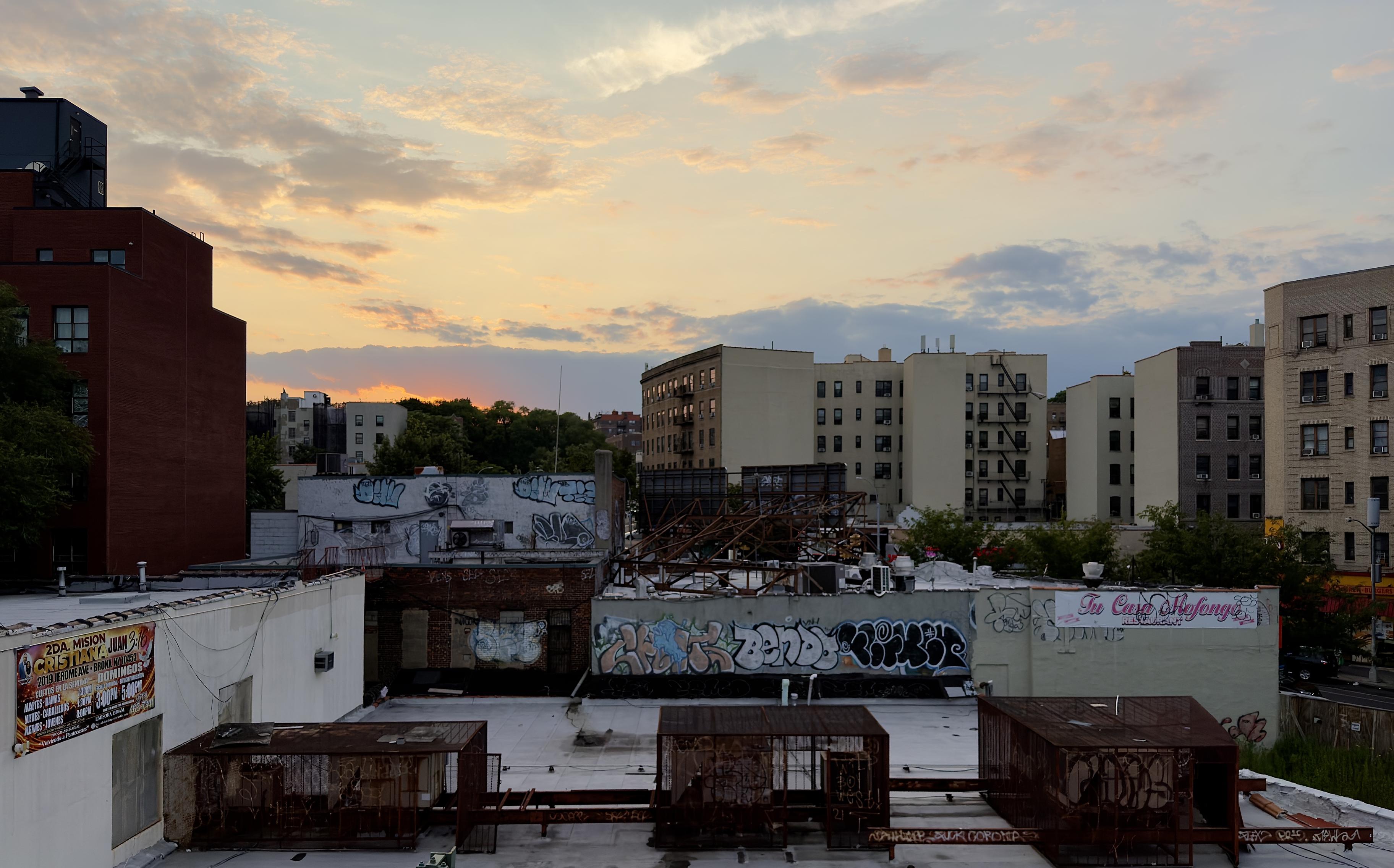 A color, wide-angle photograph in landscape orientation that captures a sunset over the Morris Heights neighborhood of the Bronx, New York, framed by various urban structures. In the foreground, the flat, grey roof of a low-rise commercial building features three rusty, rectangular metal cage-like structures housing HVAC units, aligned horizontally, with the tops of more buildings visible below. A tall, dark red building with vertical rows of windows stands on the far left, while mid-rise apartment buildings in shades of beige and brown fill the midground, featuring multiple dark windows and exterior fire escapes. The buildings in the midground are adorned with extensive graffiti, including bold, stylized lettering and abstract designs on low walls and storefronts, along with a banner for a Spanish-language Christian event on the lower-left wall and a "Tu Casa Mofongo" restaurant sign on the right. Beyond the urban rooftops, a line of green trees marks a slight rise in the landscape, leading to a dramatic sunset sky that glows with vibrant orange and gold hues just above the horizon. Above is a mix of pale blue and grey clouds is scattered across the fading evening sky, catching the soft, lingering light of dusk.