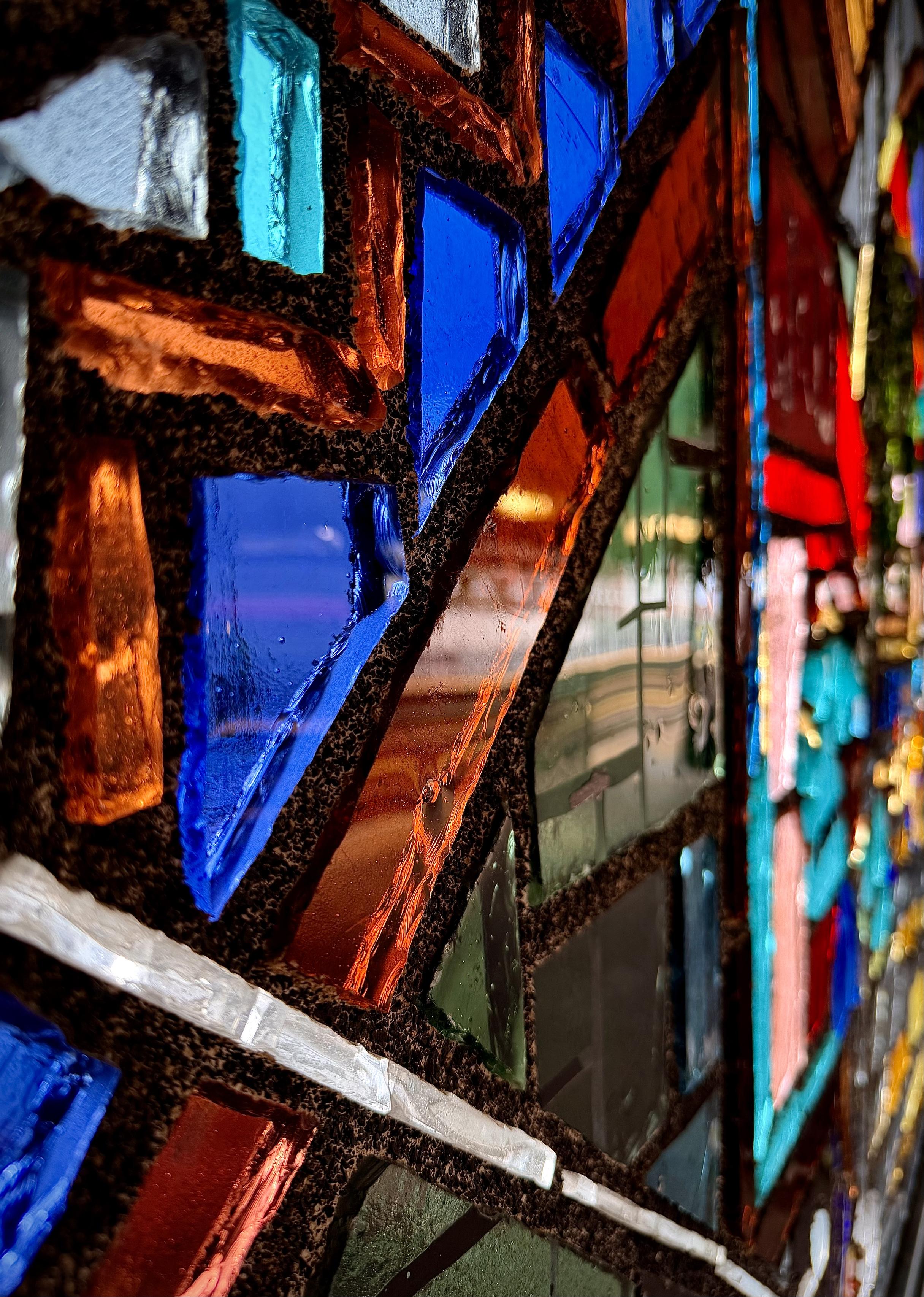 A color, medium-angle photograph in portrait orientation that captures a vibrant mosaic of stained glass at the Freeman Street subway station in the Bronx, New York City. The thick, textured chunks of glass are set into dark, gritty cement channels, creating a rough, irregular grid pattern across the frame. The perspective shifts from crisp, sharp focus in the immediate foreground to a soft blur as the glass pieces recede toward the right background. The color palette features primary and secondary shades, prominently capturing rich cobalts, deep oranges, clear whites, and muted greens. Light catches the irregular surfaces and rough edges of the individual glass pieces, creating bright glints and reflective highlights that reveal their thickness and dimension.