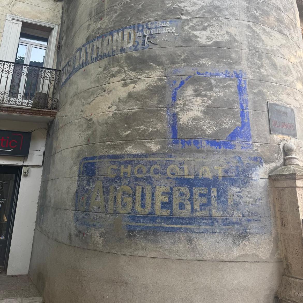 Portion of a curved, windowless, wall with a smooth patchy grey render. Parts of the wall contain fading painted signs formed of short horizontal bands, with the uppermost aligned left on the wall and the one much further below aligned right.

The lower layer of the top sign was painted in blue, while the more recent "Salon Raymond" sign is in white with a yellowy-orange block shade. The chocolate sign was once white lettering on a blue background.

Above the chocolate one is a square border, also in blue paint, but with nothing inside to indicate what, if anything, this once advertised.