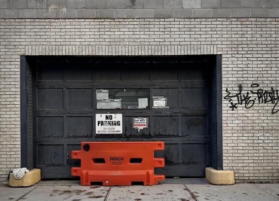 A color, wide-angle photograph in landscape orientation that captures a dark, paneled garage door recessed within a light gray brick wall along Sedgwick Avenue in the Bronx, New York City on a clear Summer morning. The garage door features a multi-paneled design painted in a dark, weathered charcoal color, with several small rectangular notices taped across its upper panels and a larger white "NO PARKING 24 HOUR ACTIVE DRIVEWAY" sign affixed to the lower left. Just above that sign, a smaller white "DO NOT TRESPASS" sticker is visible. The surrounding wall is built from light-colored, textured rectangular bricks, with a band of plain gray concrete blocks forming the upper edge of the structure. To the right of the garage entrance, black stylized graffiti is scrawled across the brick surface. Positioned directly in front of the garage door is a bright orange, interlocking plastic safety barrier. The lighting is even and diffused, highlighting the urban textures of the brickwork, weathered paint, and pavement.