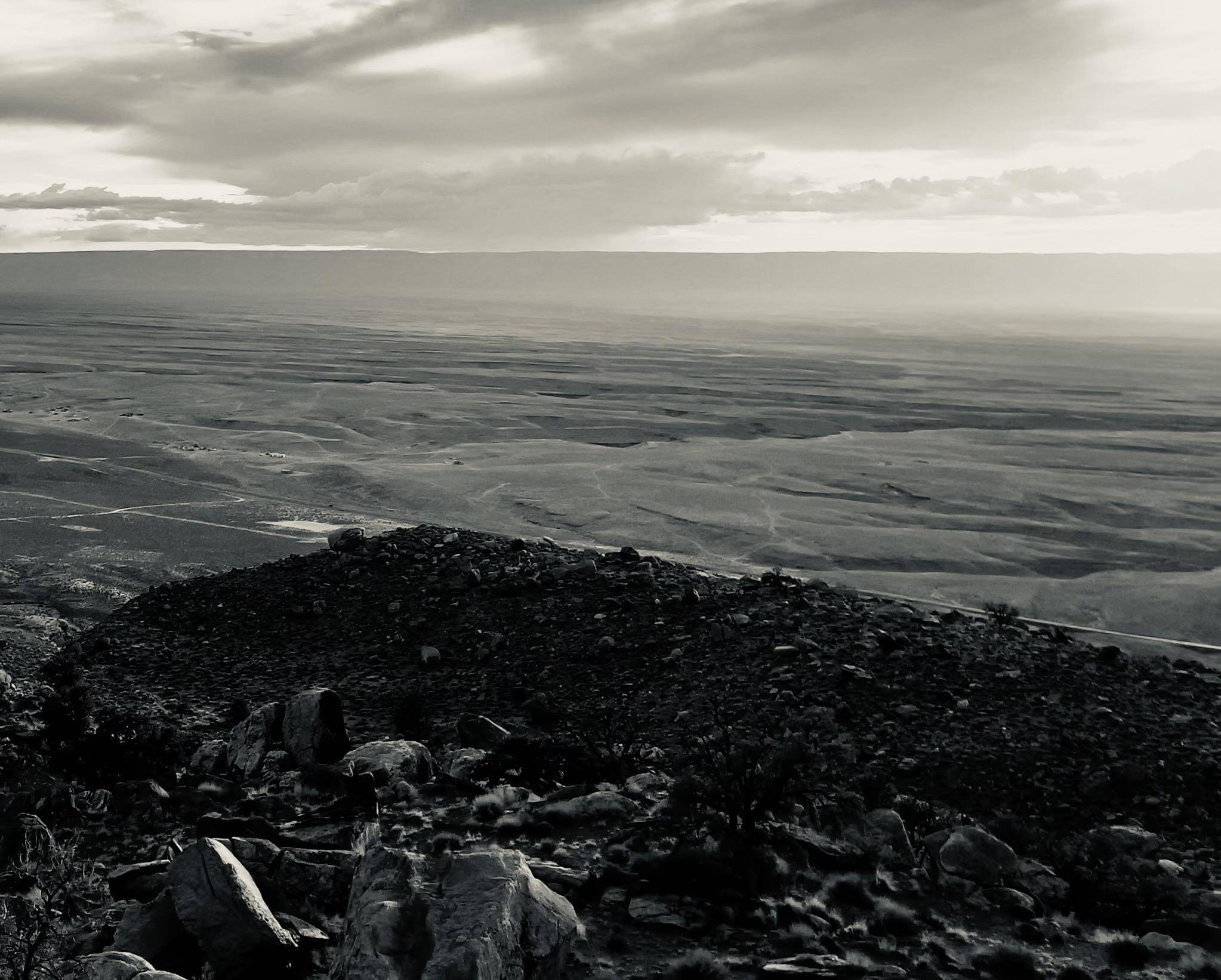 A noir, wide-angle photograph landscape orientation that captures an expansive, elevated view of Marble Canyon, Arizona, under a dramatic, heavily clouded sky. In the foreground, a dark, rocky outcropping is strewn with jagged boulders and sparse desert vegetation, rendered in deep, shadowed tones. The terrain slopes downward into a vast, sweeping plateau marked by subtle parallel ridges, faint dirt tracks, and wide-open arid spaces that recede toward a distant horizon. Above the flat, expansive desert floor, a thick layer of layered storm clouds stretches across the upper portion of the frame, diffusing a moody, directional light. Breaks in the cloud cover create bright horizontal bands of light on the far horizon, contrasting sharply with the somber, cinematic tone of the scene.