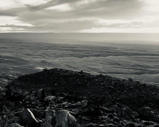 A noir, wide-angle photograph landscape orientation that captures an expansive, elevated view of Marble Canyon, Arizona, under a dramatic, heavily clouded sky. In the foreground, a dark, rocky outcropping is strewn with jagged boulders and sparse desert vegetation, rendered in deep, shadowed tones. The terrain slopes downward into a vast, sweeping plateau marked by subtle parallel ridges, faint dirt tracks, and wide-open arid spaces that recede toward a distant horizon. Above the flat, expansive desert floor, a thick layer of layered storm clouds stretches across the upper portion of the frame, diffusing a moody, directional light. Breaks in the cloud cover create bright horizontal bands of light on the far horizon, contrasting sharply with the somber, cinematic tone of the scene.