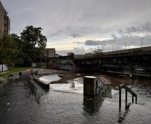 A color, wide-angle photograph in square aspect ratio that captures the River Avenue skate park in the Bronx on an overcast morning between rainstorms. The sky overhead is heavy with layered grey clouds, though a pale, muted band of warm light breaks through low on the horizon behind a line of distant buildings. In the foreground, slick, dark grey brick paving gleams with pooled rainwater, reflecting the gloomy atmosphere. A short flight of steps with black metal handrails leads down from the right foreground, while concrete ledges, banks, and ramps curve through the central skate area, partially submerged under large sheets of murky water. In the mid-ground, vibrant layers of graffiti cover the concrete retaining walls and the massive pillars supporting an elevated subway train line structure that spans the right side of the scene. Lush green trees stand tall on the left, contrasting against the industrial concrete and the weathered brick apartment buildings rising further in the background. The overall mood is quiet and desolate, highlighting the aftermath of a downpour in an urban landscape.