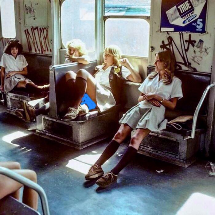 A color, wide-angle photograph in square aspect ratio that captures the interior of a graffiti-covered New York City subway car, featuring several school-aged girls in uniform sitting on brown benches. Sunlight streams in through the large windows, illuminating the metallic structure, the dark floor, and the passengers. On the right, a blonde girl in a white short-sleeve shirt, a light-colored pleated skirt, knee-high dark socks, and brown shoes relaxes with her legs stretched forward toward the foreground, her body angled slightly back against the seat. Next to her in the middle section, another blonde girl wearing a vest and light skirt leans against the window frame, with a smaller child peeking up from behind the seat divider. Further down the car to the left, another student sits against the wall beneath heavy red graffiti scrawled across the interior paneling. The entire background is marked by weathered transit details, including metallic handles, scratched surfaces, and a faded cigarette advertisement reading "TRUE" affixed to the wall near the top right corner. The lighting casts distinct shadows across the floor.