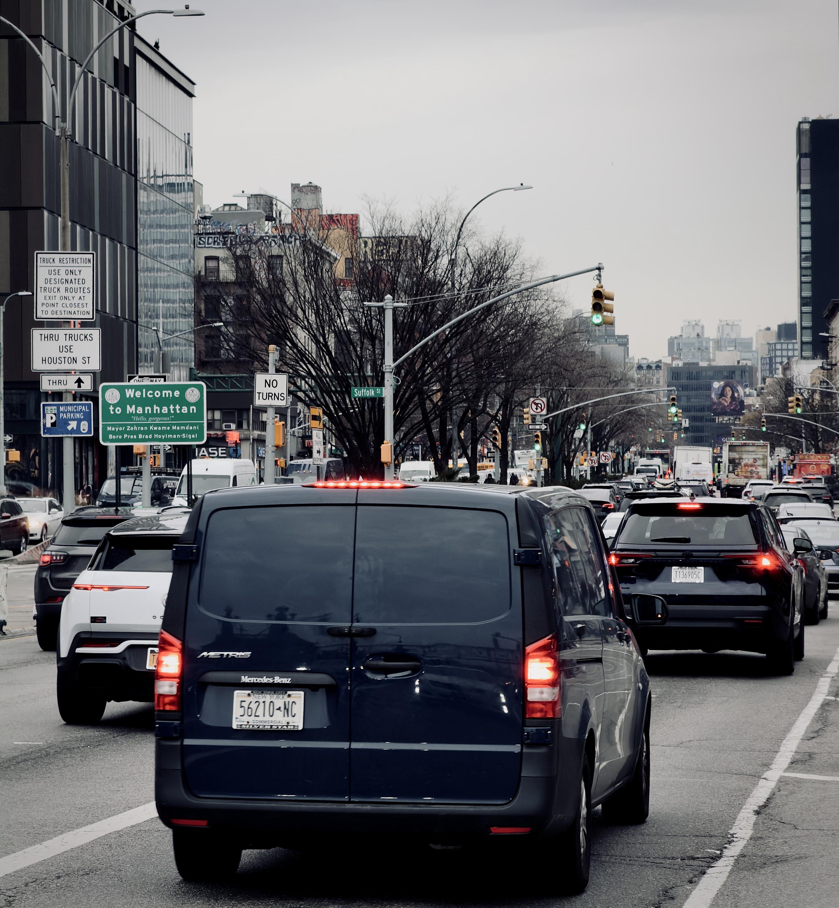 A color, wide angle photograph in square aspect ration capturing a daytime view facing down a wide multi-lane street in Manhattan under a uniform gray, overcast winter time sky. The shot is tightly framed by heavy, slow-moving urban traffic captured from behind a dark blue Mercedes-Benz Metris cargo van in the center foreground, its rear license plate visible and taillights illuminated red. Flanking the central van are several other vehicles, including a white SUV to its left and a dark SUV to its right, with a dense queue of cars, trucks, and vans extending into the hazy distance under a complex network of traffic lights, streetlamps, and overhead green and white street signs. To the left, a tall modern building with dark, paneled vertical windows rises beside older, graffiti-tagged brick architecture further down the block. A large green street sign prominently reads "Welcome to Manhattan" alongside regulatory parking and truck restriction panels mounted on poles along the sidewalk. The background is dense with bare deciduous trees lining the street, interspersed with low-rise and high-rise apartment buildings under a flat, pale horizon line.