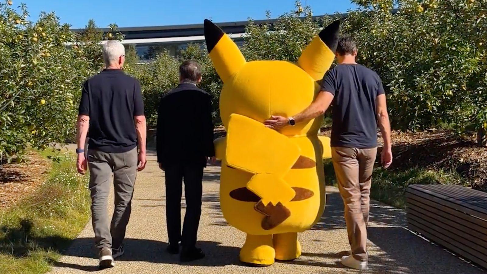 A color, wide-angle photograph taken outdoors within the Apple Park campus during the daytime. Facing away from the camera while walking, Tim Cook, a representative from the Pokémon team, a person dressed in a large Pikachu costume, and John Ternus. Ternus has his left arm around Pikachu’s shoulder.
