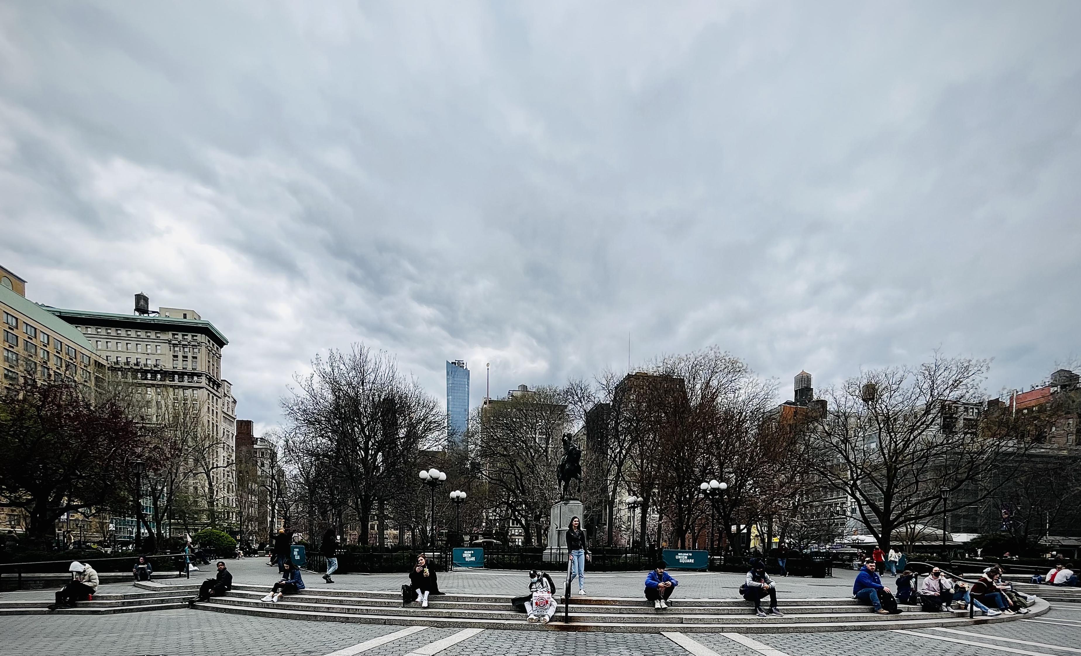 A color, wide-angle photograph in landscape orientation that captures a wide, curving set of stone steps and plaza area at Union Square Park in Manhattan under an expansive, heavily overcast sky. The gray clouds dominate the upper half of the frame, diffusing the afternoon light across the urban landscape. In the midground, several individuals are scattered along the multi-tiered stone steps. A central equestrian statue stands surrounded by leafless, early-spring deciduous trees, while historic masonry buildings flank the left side of the plaza and a tall, modern glass skyscraper pierces the skyline in the distance. The people in the plaza are dressed in warm winter or transitional spring layers, including dark jackets, hoodies, and jeans. Most figures are sitting individually or in small, isolated pairs on the lower risers of the curved staircase, maintaining physical distance. Low green park signage barriers are visible near the base of the trees, and the overall atmosphere is subdued, calm, and spacious under the heavy cloud cover.