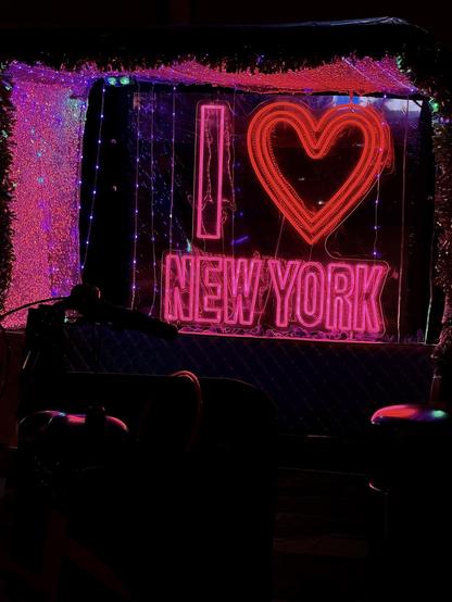 A color, wide-angle photograph taken along Madison Avenue in Manhattan at night, capturing a bicycle taxi highlights a glowing pink neon sign that reads "I ❤️ NEW YORK" mounted against the back partition. The sign's bright pink and red tubes illuminate the interior, surrounded by a curtain of vertical fairy lights and shimmering tinsel lining the overhead roof and side panels. In the foreground, the dark silhouette of the bicycle's handlebars, seat, and structural frame is visible against the neon glow. The overall atmosphere is dark, saturated with vibrant pink and purple ambient light reflecting off the metallic and quilted surfaces of the cab's interior.