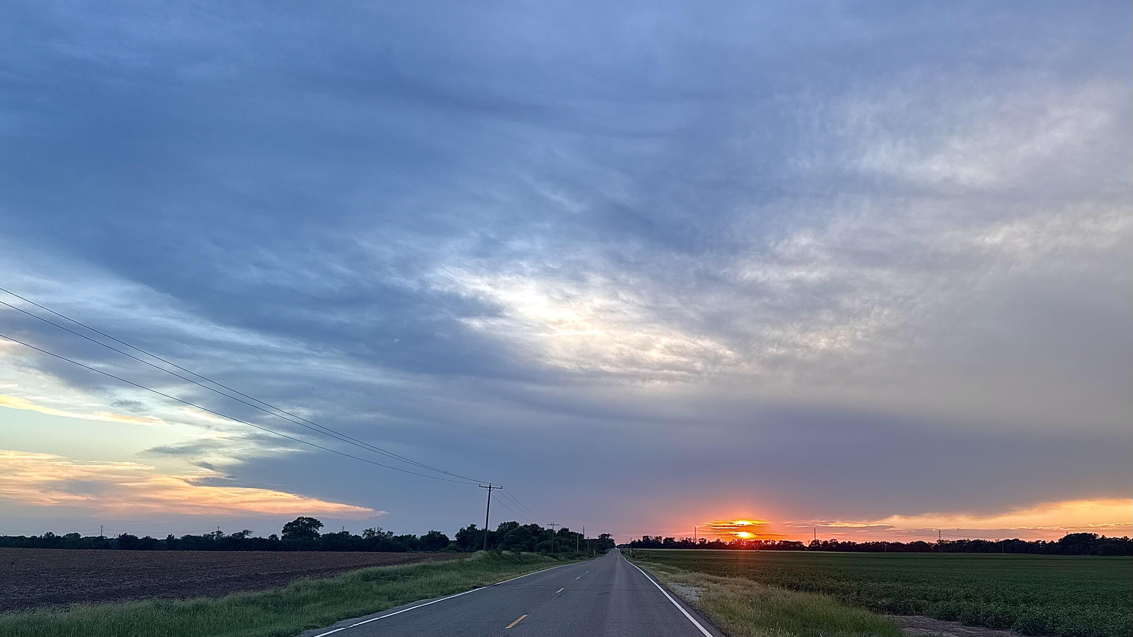 A color, wide-angle photograph in landscape orientation that capture a serene sunset outside of Dodge City, Kansas. A straight two-lane country highway cuts vertically through the center of the frame, receding toward a distant horizon under a vast, dramatic sky. The asphalt road features a dashed yellow centerline and white edge lines, flanked by grassy shoulders, flat agricultural fields, and low treelines stretching across the horizon. On the left side of the road, dark plowed earth contrasts with a strip of green grass, while the right side features a lush green crop field. Utility poles with thin power lines stand vertically on the left side, tilting slightly against the landscape.The upper portion of the frame is dominated by a sweeping expanse of cool blue and gray clouds that stretch across the sky in layered, wispy textures. Near the lower right horizon, the low sun dips below the tree line, creating a vibrant sliver of intense orange and yellow light that peeks out just above the flat terrain. To the far left, a gentle gradient of pale yellow and orange sunset light catches the lower edge of the cloud deck, casting a quiet, luminous glow over the rural horizon.