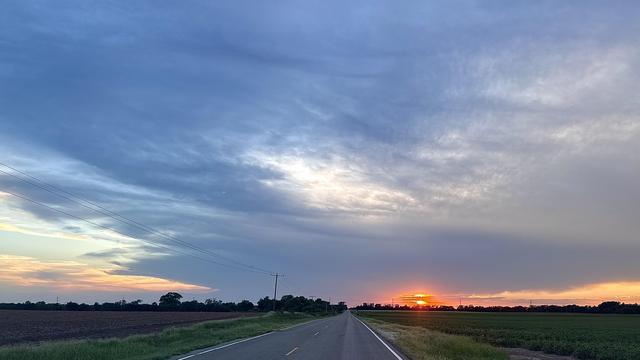 A color, wide-angle photograph in landscape orientation that capture a serene sunset outside of Dodge City, Kansas. A straight two-lane country highway cuts vertically through the center of the frame, receding toward a distant horizon under a vast, dramatic sky. The asphalt road features a dashed yellow centerline and white edge lines, flanked by grassy shoulders, flat agricultural fields, and low treelines stretching across the horizon. On the left side of the road, dark plowed earth contrasts with a strip of green grass, while the right side features a lush green crop field. Utility poles with thin power lines stand vertically on the left side, tilting slightly against the landscape.The upper portion of the frame is dominated by a sweeping expanse of cool blue and gray clouds that stretch across the sky in layered, wispy textures. Near the lower right horizon, the low sun dips below the tree line, creating a vibrant sliver of intense orange and yellow light that peeks out just above the flat terrain. To the far left, a gentle gradient of pale yellow and orange sunset light catches the lower edge of the cloud deck, casting a quiet, luminous glow over the rural horizon.
