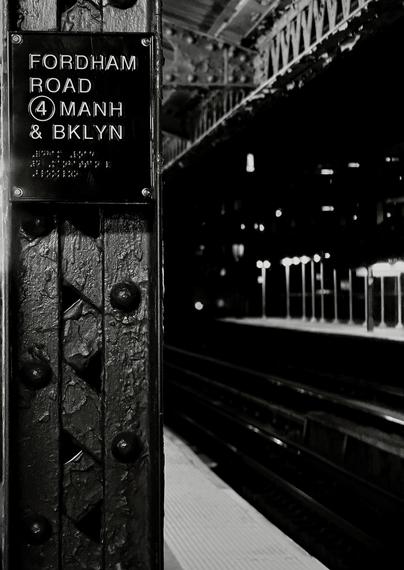 A noir, wide-angle photograph in portrait orientation that captures an elevated subway platform at night, viewed from a tight perspective framing a heavy, weathered structural column in the immediate foreground. The dark, textured iron beam is heavily bolted and shows layers of thick, chipped paint, with a rectangular station sign affixed near the top. The sign features raised white sans-serif text reading "FORDHAM ROAD," a circled number "4," and "MANH & BKLYN," accompanied by a block of tactile Braille lettering underneath. Bright glints of light catch the edges of the metal lettering and the screw heads securing the plaque, emphasizing the industrial texture of the column. In the blurred background, the dark empty tracks stretch into the distance alongside the edge of the dimly lit platform. Across the tracks, a series of glowing vertical light fixtures or poles stand in a row against the pitch-black night, casting a soft, diffuse glow on an opposing platform structure. Overhead, intricate steel girders and trusses of the elevated station roof vanish into deep shadow, creating a moody, cinematic atmosphere dominated by stark contrasts between deep blacks and brilliant highlights.