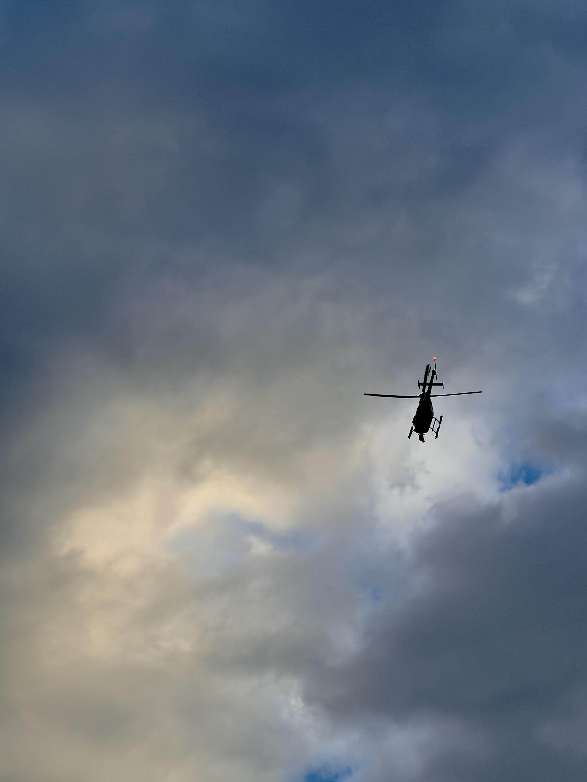A color, wide-angle, low-angle photograph in portrait orientation that captures the dark silhouette of a helicopter flying through a dramatic, cloudy sky illuminated by the soft, golden glow of sunrise. The helicopter is positioned toward the lower right of the frame, angled slightly upward with its main rotor blades spread horizontally and a small red light blinking on its tail assembly. The vast expanse above and around the aircraft is filled with dense, billowing gray and blue clouds, while a break of warm, pale yellow light softly brightens the lower left area of the dramatic morning scene.